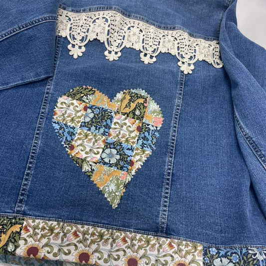 A blue denim jacket laid flat, featuring a large heart-shaped patchwork floral design on the back, a cream lace band near the collar, and a decorative floral border along the hem.